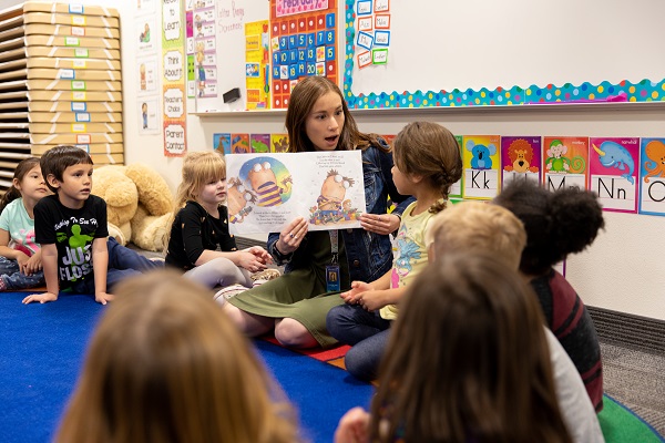 A teacher sitting in a classroom on the ground with preschool students is reading a book to them