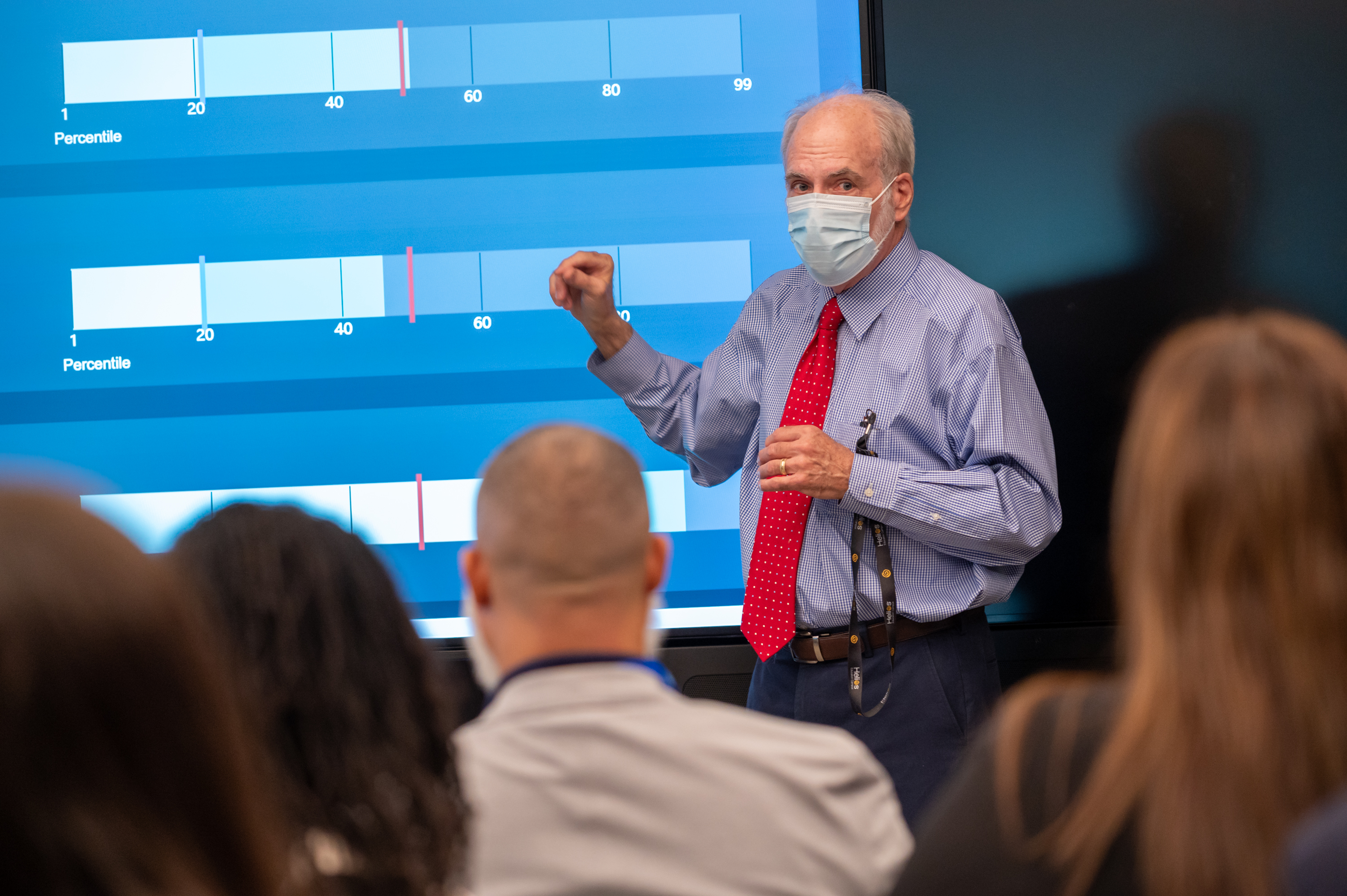 Joseph O'Reilly wearing face mask stands in front of large blue screen presenting data to a group of adults.