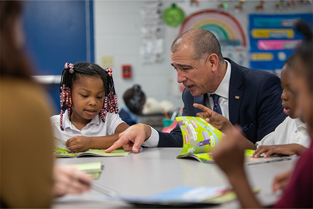 Paul Luna sitting at desk in classroom with two elementary school children.